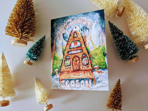 Christmas Card of an A-Frame Cabin with Christmas lights in the Snow surrounded by Watercolor Galaxy and Trees that says "Coziest Season"