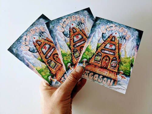 Christmas Card of an A-Frame Cabin with Christmas lights in the Snow surrounded by Watercolor Galaxy and Trees that says "Coziest Season"