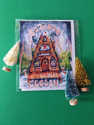 Christmas Card of an A-Frame Cabin with Christmas lights in the Snow surrounded by Watercolor Galaxy and Trees that says "Coziest Season"